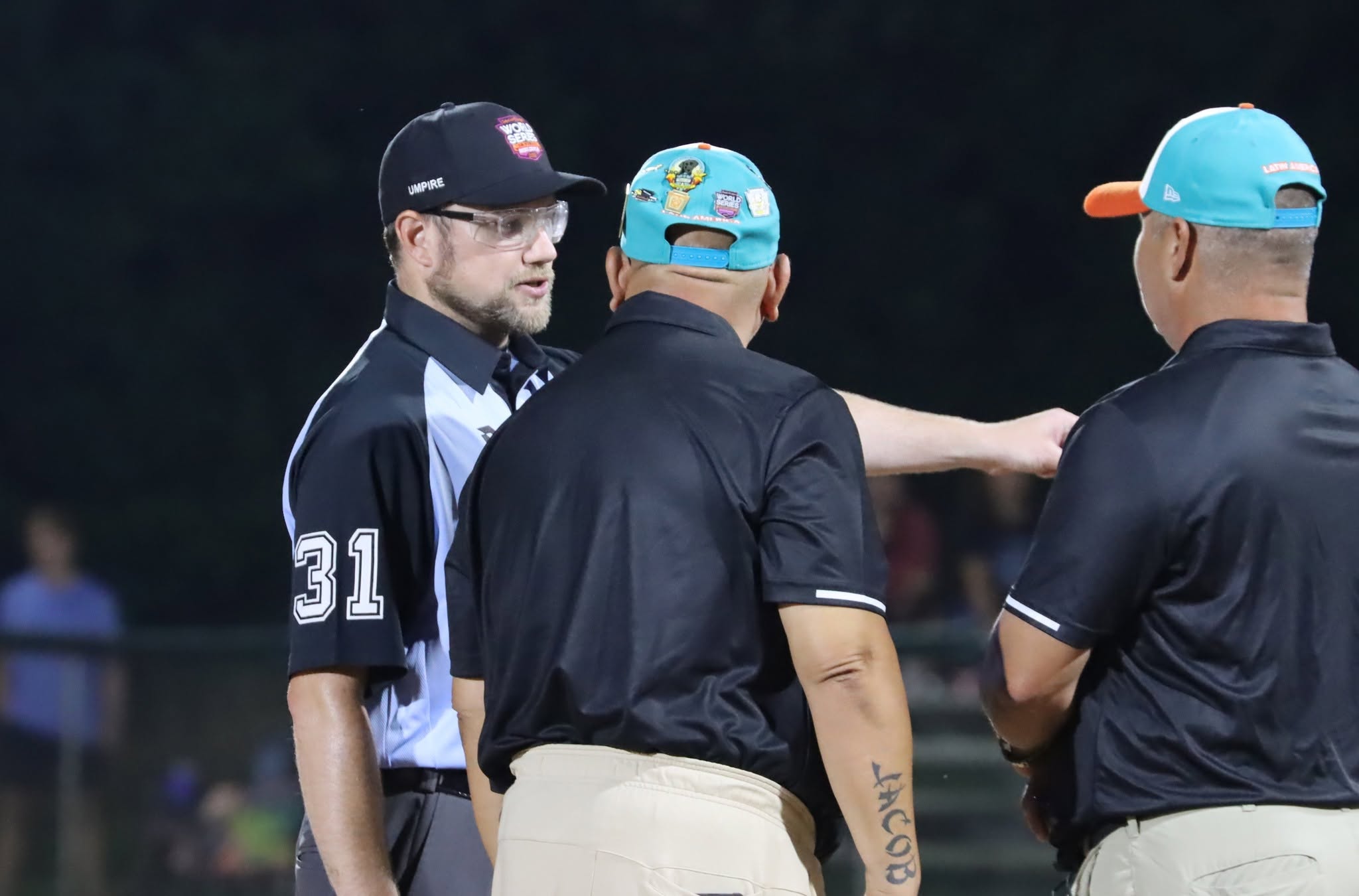 Gareth Gilson in umpire uniform, number 31, standing between two coaches mid-conversation during a night game.